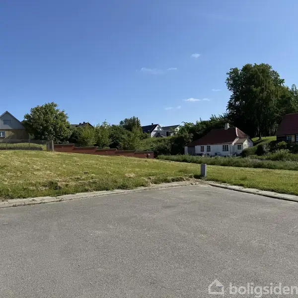 En tom byggegrund ligger stille. Den er omgivet af grønne græsplæner og forskellige huse i baggrunden under en blå himmel.