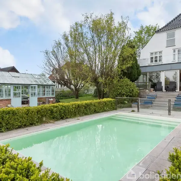 En swimmingpool er omgivet af dekorative buske, mens et stort hus med træer i baggrunden har åbne dobbeltdøre. Et drivhus ses til venstre for husets have.
