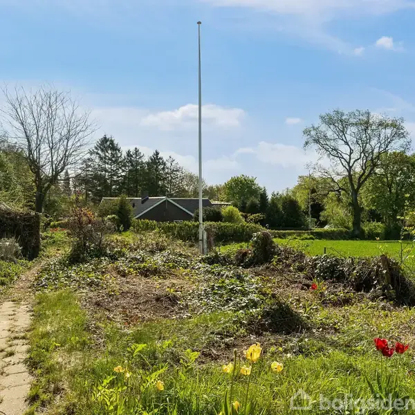 En flagstang står i en have med vilde blomster og stier. I baggrunden ses et hus, marker og træer under en klar blå himmel.