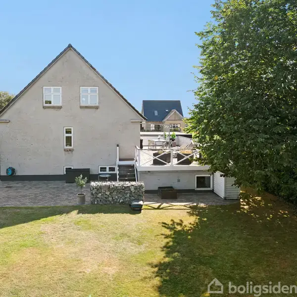 Et gråhvidt hus med to etager står med en stor terrasse foran. En grøn have omkranser bygningen, hvoraf en stor træ sørger for skygge til venstre.