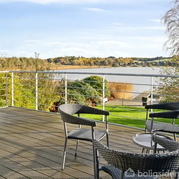Terrassemøbler står på en træterrasse med udsigt over en sø. Omkring er grønne træer, og himlen er blå. Det er en rolig, naturskøn scene i et rekreativt udemiljø.