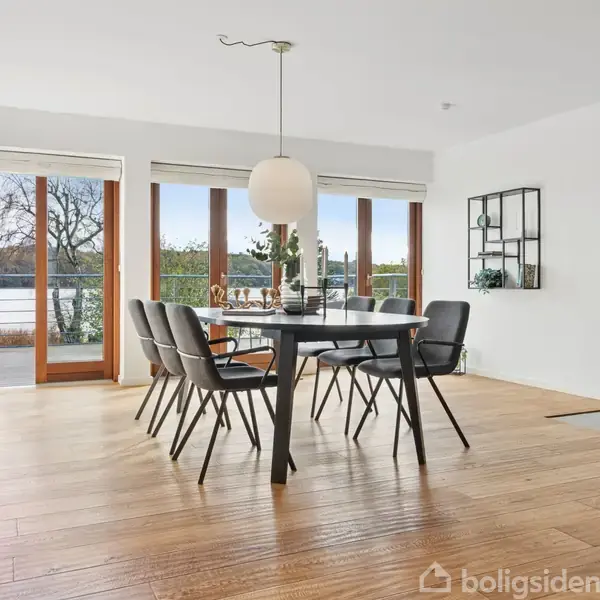 Et spisebord med seks stole står i et lyst rum med trægulv og store vinduer, der åbner til en terrasse med udsigt. En lampe hænger over bordet.