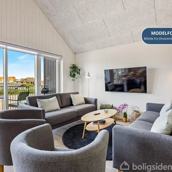 Stue med grå sofaer og lænestole, der står rundt om et rundt sofabord. Et vægmonteret TV viser en udsigt. Store vinduer åbner mod en balkon. Tekst: "MODELFOTO Billede fra tilsvarende lejlighed".