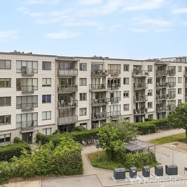 En moderne lejlighedsbygning med flere etager, hvor balkoner vender ud mod en grøn gård. Foran bygningen er der parkeringspladser og affaldscontainere.
