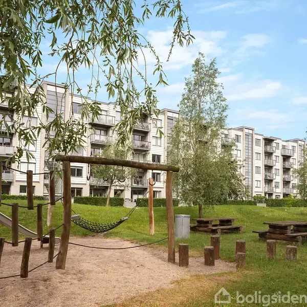 Legeplads med klatrestativer står på græsområde foran en række lejlighedsbygninger. Området inkluderer træborde og bænke, omkranset af træer under en blå himmel.