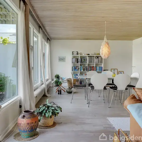 Stue med spisebord og seks hvide stole, omgivet af store vinduer og planter. En reol fyldt med bøger står op ad væggen, og en unik lampe hænger over bordet.