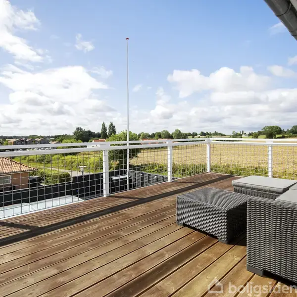 Terrasse med grå sofa og bord står på en træbelægning. Udsigt til marker, huse og en blå himmel med spredte skyer i baggrunden.
