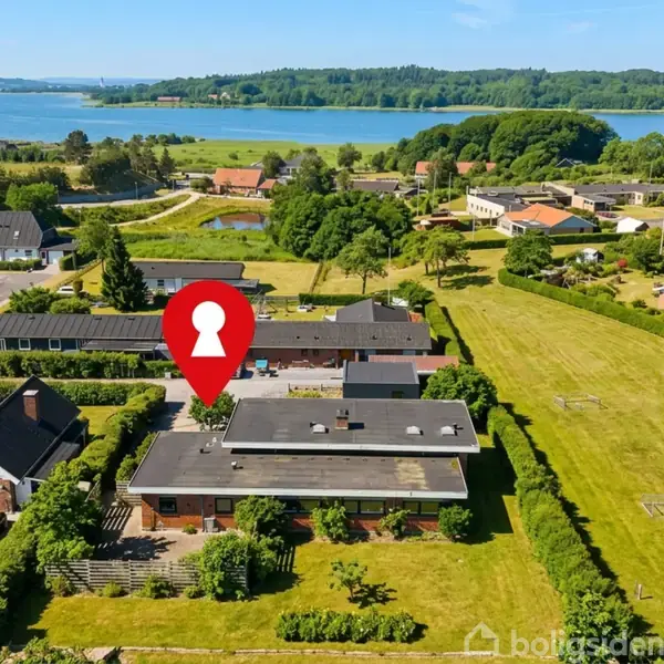 En enetages bygning markeret med en rød nøgleformet placeringstegn, omgivet af grønt græs og flere huse i et landligt område ved en sø.