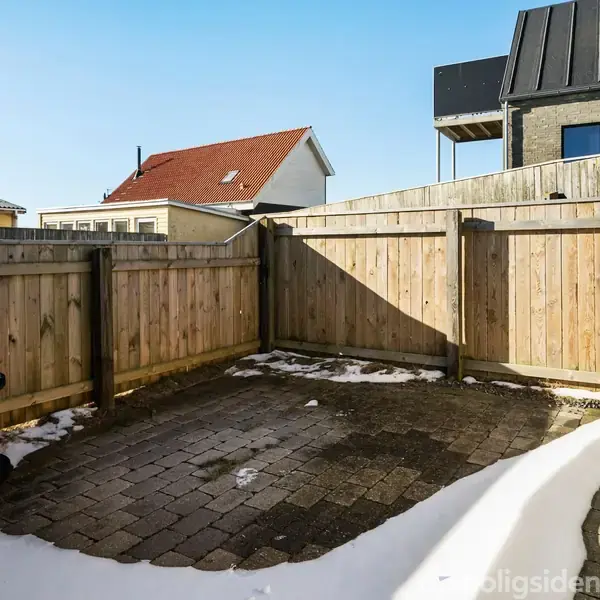En lille flisebelagt gårdhave omringet af et træhegn. Sne ligger langs kanterne. Omkring gårdhaven ses dele af nærliggende hustage under en klar blå himmel.