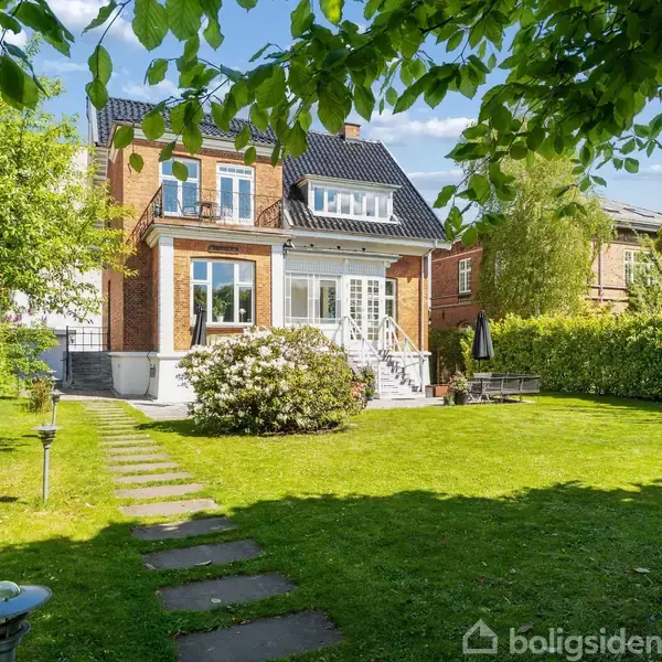 Et rødt murstenshus med sort tag står i en frodig have. Træer og en plæne omkranser huset, og der er en sti af fliser, der fører til en terrasse.