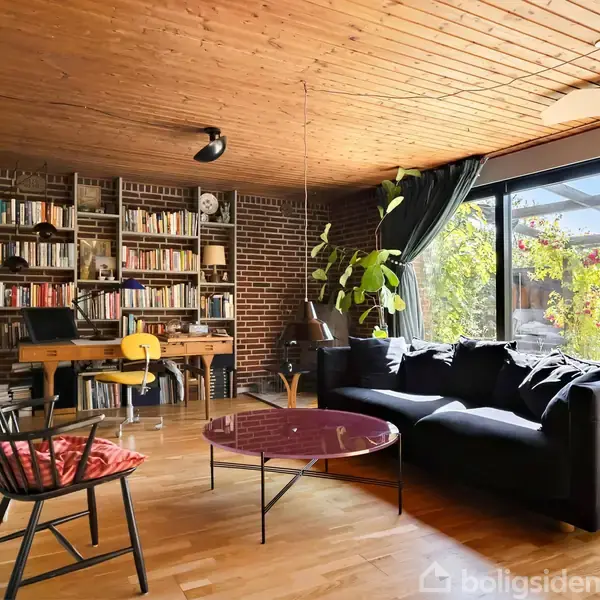 En stue med en sort sofa, et rundt bord og en reol fyldt med bøger. Store glasvinduer til højre giver udsigt til en have med grønne planter.