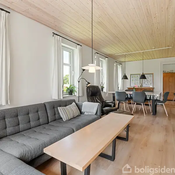 Stue med grå sofa står ved et stort vindue, der lader lys strømme ind. Bagved ses spisebord med stole. Rummet har træloft og minimalistisk indretning.
