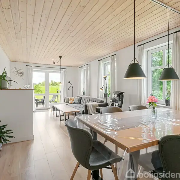 Spisebord dækket med plastikdug, omgivet af stole, står i et lyst rum. I baggrunden ses en stue med sofa og vinduer med udsigt til en grøn have.