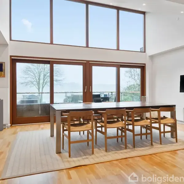 Et stort spisebord med otte træstole står på et tæppe i en lys stue. Rummet har store vinduer, der giver udsigt til en terrasse og natur udenfor.