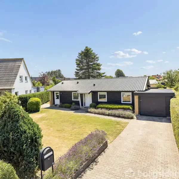 Et hus med en garage står i en velplejet have med en asfalteret indkørsel og grønne hække. Naboen har et lignende hus i baggrunden under en klar blå himmel.