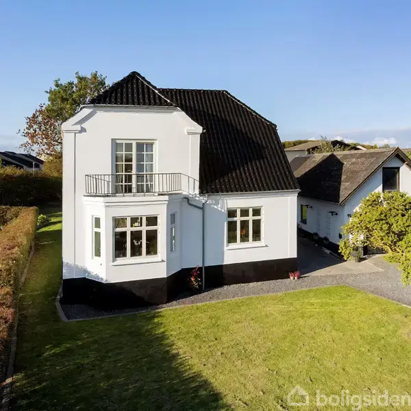 Et hvidt hus med sort tag står i en velplejet have. Huset har en mindre balkon og er omgivet af hække og træer i et beboelsesområde.