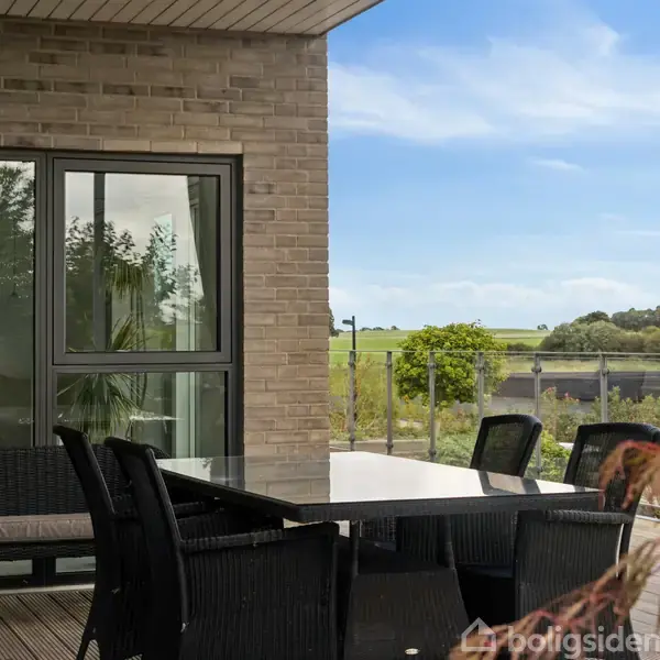 Terrasse med sort havemøbelgruppe står foran en murstensvæg med vinduer. I baggrunden ses en grøn udsigt med en blå himmel.