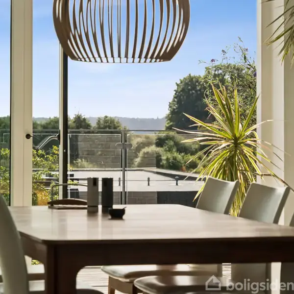 Et spisebord med hvide stole står under en moderne, træstruktur loftlampe. Udenfor er der en terrasse og udsigt over et grønt landskab med træer og blå himmel.