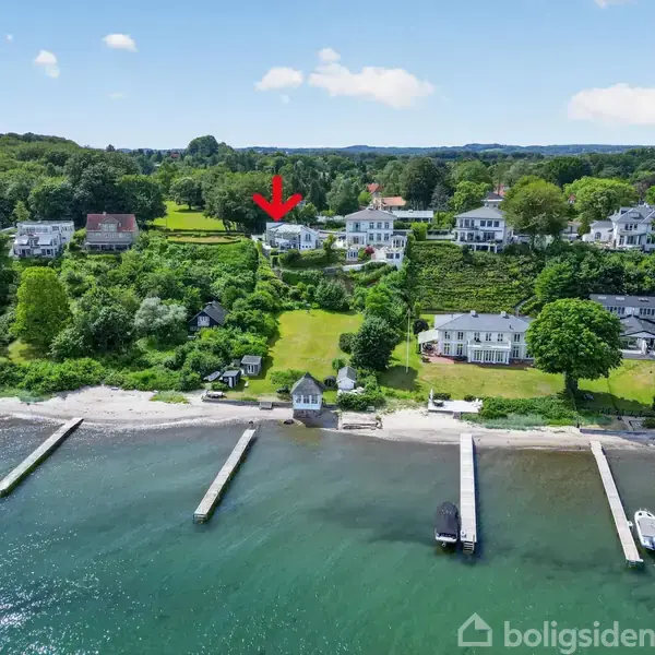 Hus markeret med rød pil ligger i grønne omgivelser nær kysten. Flere bådebroer strækker sig fra stranden ud i det klare vand. Skovklædt bakkelandskab bagved.