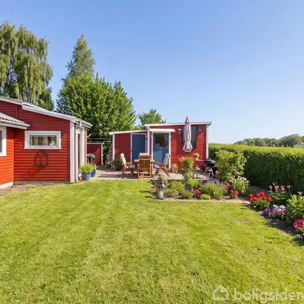 Et rødt hus med hvide karme står ved siden af en velplejet have. Et havebord med stole står på terrassen, omgivet af farverige blomster og grønne buske.