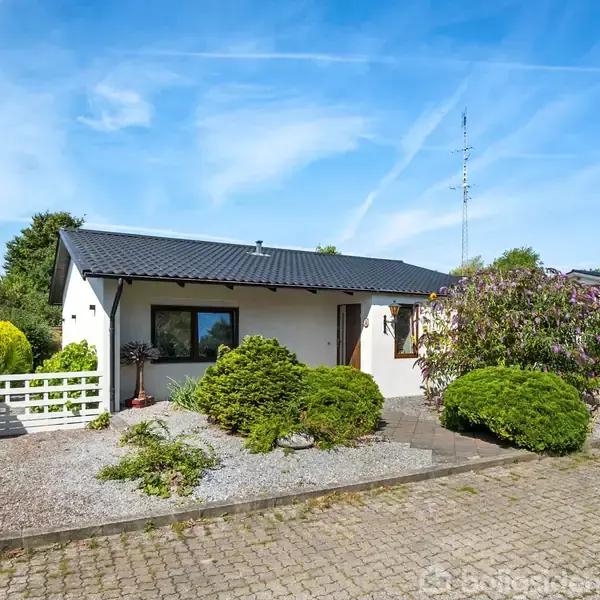 En hvid enetages villa med sort tag står på en velplejet grund. Foran huset er der buske og en flisebelagt indkørsel. Blå himmel og grønne træer omkranser scenen.