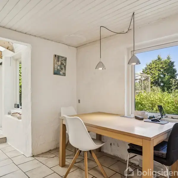 Et lyst værelse med et træbord og to stole, hvoraf én er hvid. Store vinduer giver udsigt til en grøn have. Loftslamper hænger over bordet.