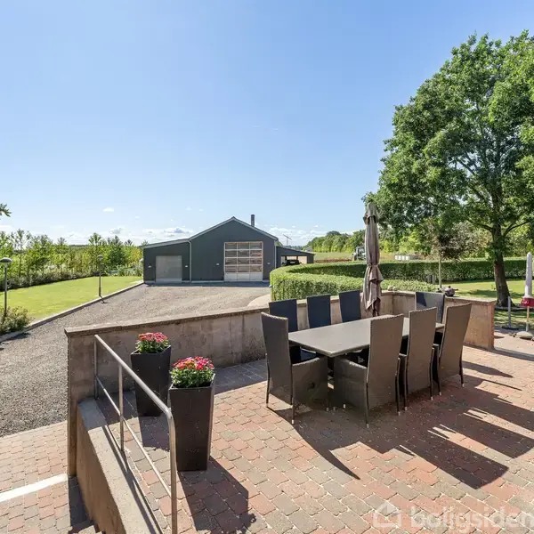 En terrasse med et bord og stole står i en have omkranset af grønne træer. I baggrunden ses en garagebygning. Solrigt vejr med blå himmel og få skyer.