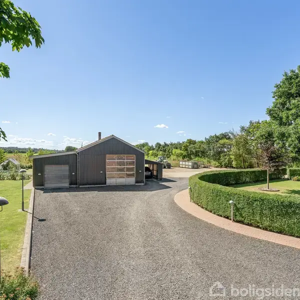 En moderne, mørk garagebygning står placeret i et åbent område. En grusvej fører op til bygningen, omgivet af velplejede grønne områder og træer under en klar blå himmel.