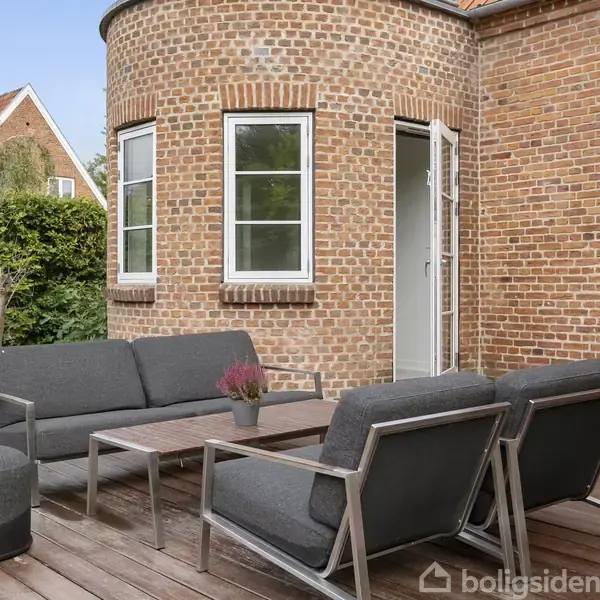 Terrassemøbler er placeret på en træterrasse foran en rund murstensbygning med vinduer. En sofa, to stole og et bord står ved huset, omgivet af grønne buske og træer i baggrunden.