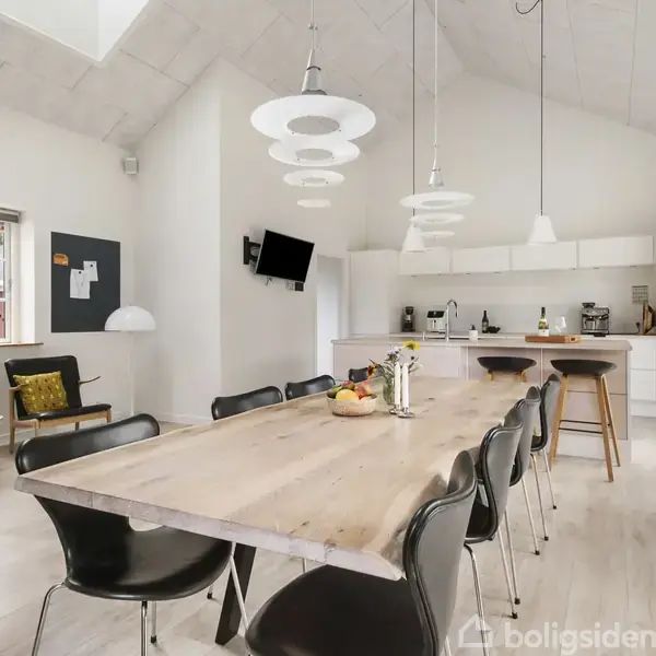 Spisebord er centralt placeret med sorte stole omkring, under moderne lamper. Køkkenet bagved har hvide skabe og indbyggede apparater. Et vindue viser en have, og en væg-TV hænger ved siden af.