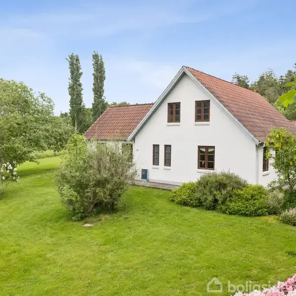 Hvidt hus med rødt tegltag, omgivet af en grøn have med buske og træer. Rolig landlig scene med åbne græsarealer og få høje træer i baggrunden. Ingen tekst fundet.
