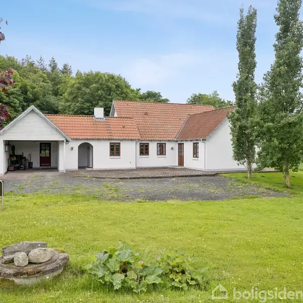 Hus med rødt tegltag og hvid facade omgivet af grøn plæne og træer. Stenbelagt indkørsel fører til carport. Blå himmel og frodig vegetation i baggrunden.