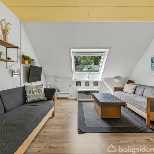 To sofaer vender mod hinanden i en hyggelig stue med skråvægge og ovenlysvindue. Trægulve, hylder med pynt og et stort sofabord i midten skaber en indbydende atmosfære.