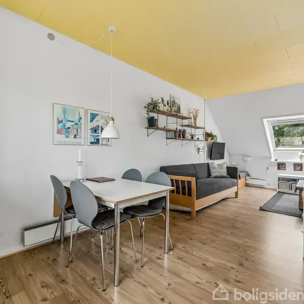 Spisebord med fire stole står på trægulv. Sofa placeret ved siden af skråvæg med stort vindue. Hylder og planter dekorerer væggen; gul loft skaber lys atmosfære.