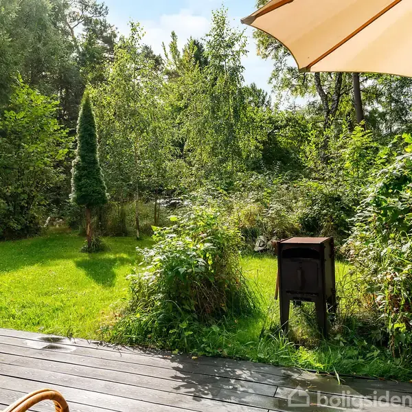 En terrassedør åbner ud mod en frodig have med grøn græsplæne og tætte buske. En fritstående brændeovn står på græsset. En parasol kaster skygge over terrassen.