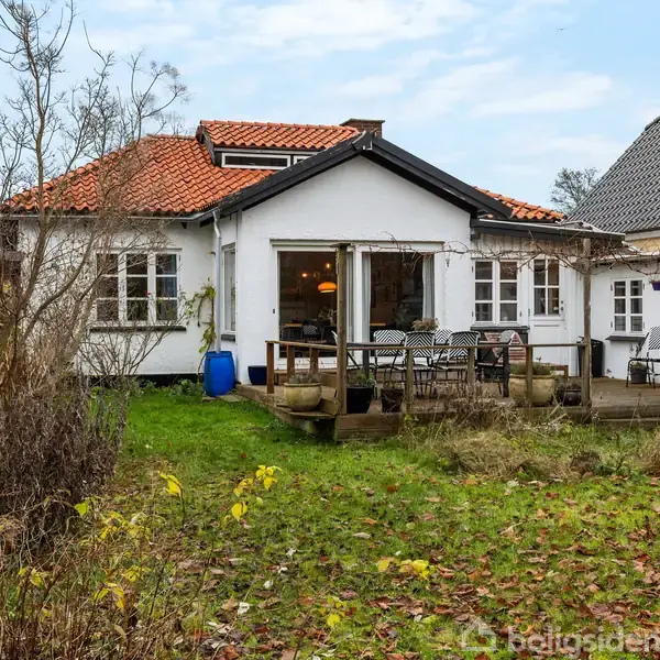 Hus med hvid facade og rødt tegltag, stående i en have med græsplæne og terrasse. Terrassen har bord og stole, omgivet af planter og buske.