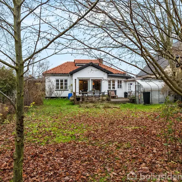 Et hvidt hus med rødt tegltag står i en have dækket af nedfaldne blade. En træterrasse med havemøbler er foran huset. Træer omkranser haven.