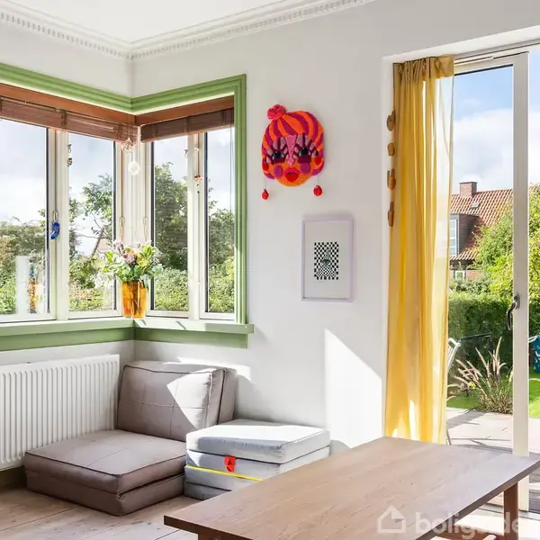 Sofa står ved vindue med grønne karme og blomster i potter, mens gul gardin rammer åbningen til en solrig have i baggrunden. En farverig dekoration hænger på væggen.