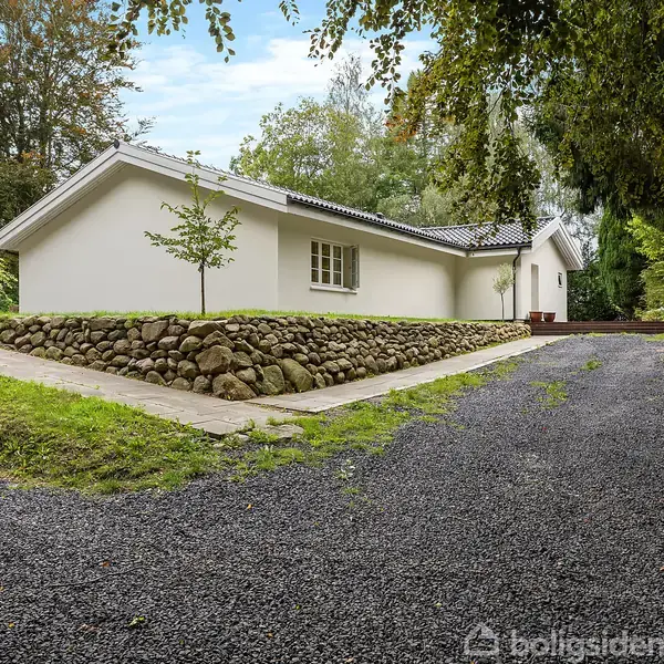 Hvidt hus står stille på en grusvej omgivet af grønne træer og buske. Huset har en mørk tagryg og en lille græsplæne foran, der er pyntet med en stengærde.