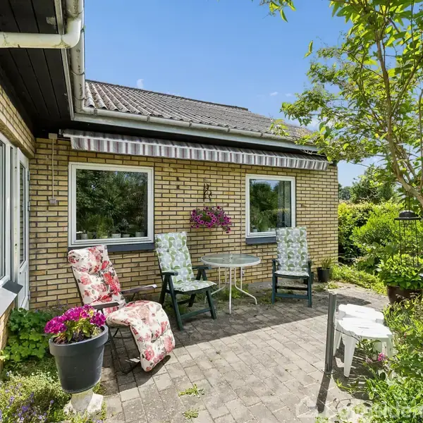 Et hyggeligt terrasseområde med to polstrede stole og et rundt bord. Det står foran et gult murstenshus med to vinduer og en markise. Omgivet af grønne planter.