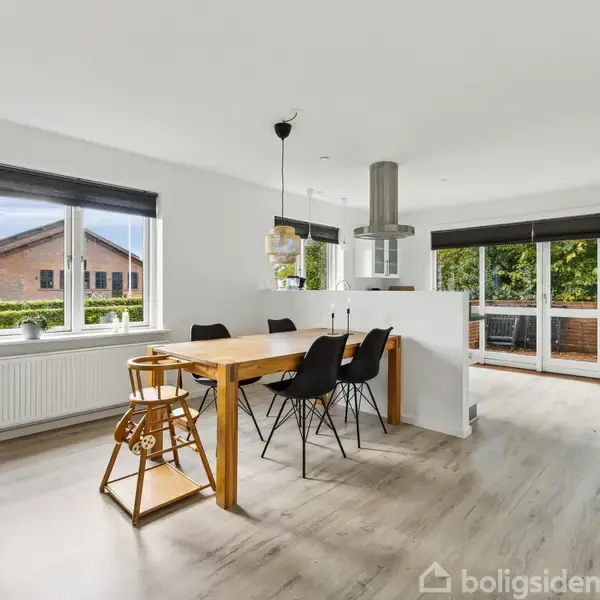 Spisebord med sorte stole står på lyst parketgulv. Et stort vindue og dør til terrasse fylder væggen, omgivet af hvidmalede vægge. En moderne lampe hænger over bordet.