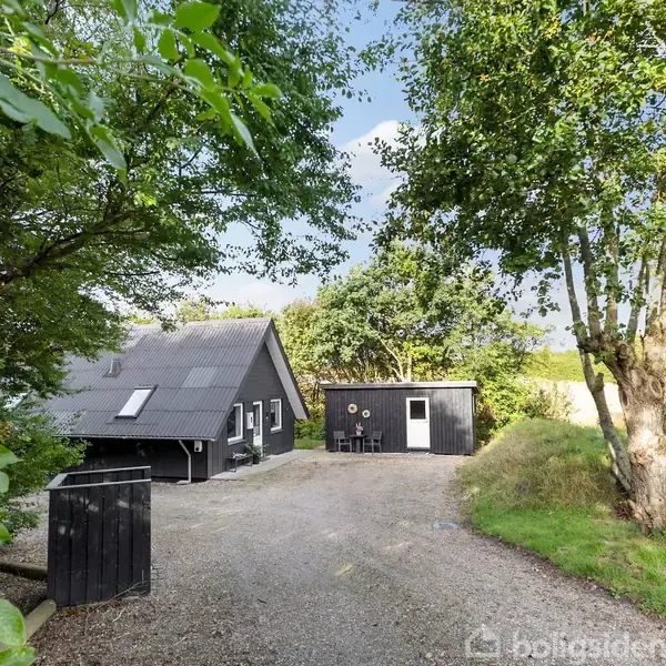 Sort træhus med skråt tag placeret i en grøn omgivelser. Der er en grusvej foran huset og træer omkring. En mindre fyrretræshytte ses tæt ved huset.