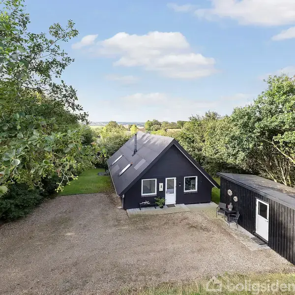 Et sorte hus ligger midt i en skovklædt have med en grusindkørsel. Et skur står ved siden af. Området er omgivet af grønne træer og lyseblå himmel.