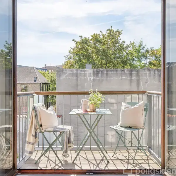 Balkon med to stole og et bord dækket med blomster og en tekande. En stribet tæppe hænger over den ene stol. Baggrunden viser et murværk og træer.