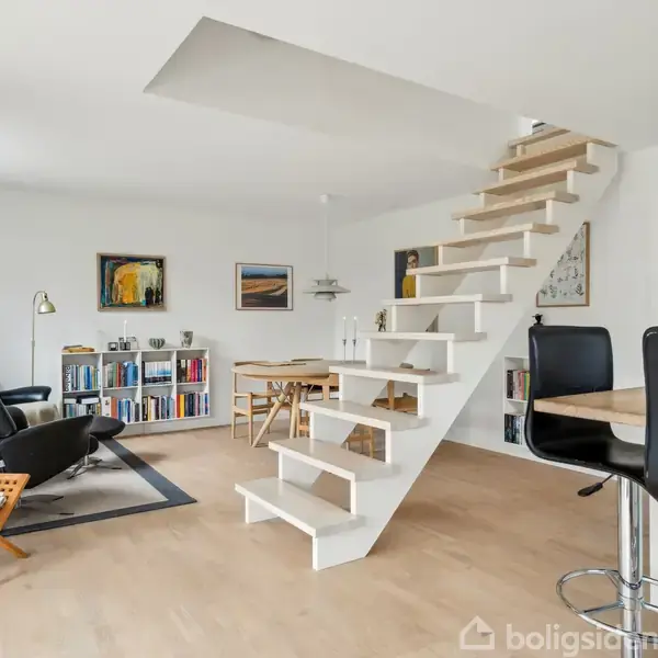 Trappe fører til anden etage i moderne opholdsområde. Omkring er lænestol, sofabord, bogreol og spisebord. Lyse vægge, træmøbler og minimalistisk indretning skaber en åben atmosfære.