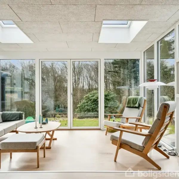 Solrig udestue med møbleret siddeplads; sofa, stole og lille bord står på lyst gulv. Store vinduer og ovenlys åbner rummet til grøn have udenfor. Ingen tekst fundet.