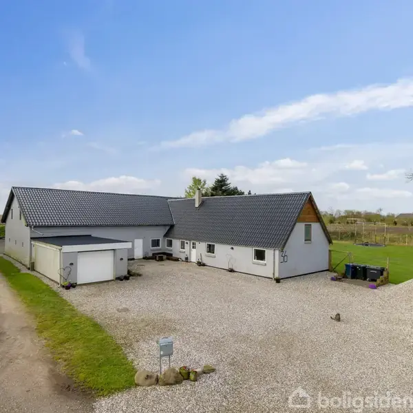 Et hvidt landhus med sort tag står på en grusbelagt gårdsplads. Vejen fører fra venstre mod huset, omgivet af grønne marker og træer under en blå himmel.