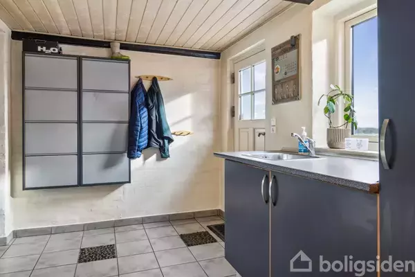 Gråt opbevaringsskab hænger på væggen, jakker hænger på kroge ved siden af. Bord med planter og rengøringsmiddel står i forgrunden. Lyse fliser på gulvet, vindue ved siden af dør.