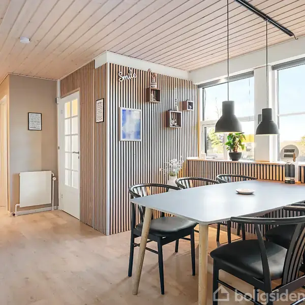 Spisebord med sorte stole står i et lyst rum med trævægge og loft. Rummet har store vinduer og tre hængende lamper over bordet. En åben køkkenindgang ses i baggrunden.