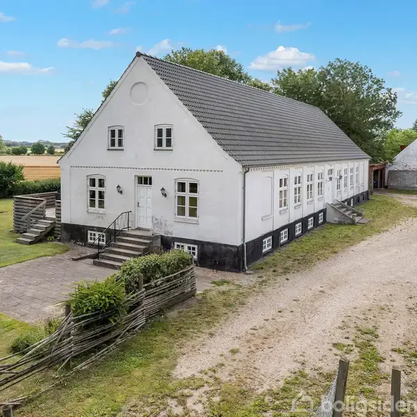 Gård bygning, stående stille, omgivet af åbne marker og blå himmel. Hvid murstensfacade med sorte detaljer og flere indgangsdøre. Grusvej fører til hovedindgangen. Kanter af grøn vegetation.